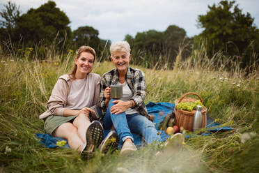 Eine glückliche ältere Mutter und eine erwachsene Tochter, die draußen in der Natur sitzen und picknicken und dabei in die Kamera schauen. - HPIF05277