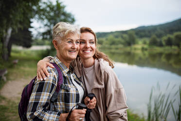 A happy senior mother hiker embracing with adult daughter when looking at lake outdoors in nature - HPIF05284