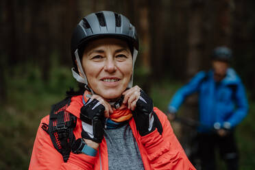 Ein älteres Radfahrerpaar, das im Herbst im Wald einen Fahrradhelm aufsetzt und in die Kamera schaut - HPIF05291