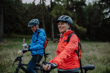 A senior couple bikers with e-bikes admiring nature outdoors in forest in autumn day. - HPIF05305
