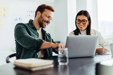 Zwei Geschäftsleute, die zusammen einen Laptop benutzen, während sie in einer Besprechung sitzen. Glückliche Geschäftsleute, die sich in einem Büro eine Folienpräsentation ansehen. - JLPSF28681