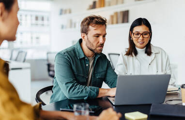 Business man using a laptop in a meeting with his team. Business professional showing his colleagues a report while sitting at a table in an office. - JLPSF28685