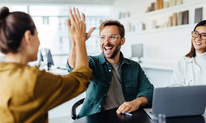 Erfolgreiche Geschäftsleute geben sich in einer Sitzung gegenseitig ein High Five. Zwei junge Geschäftsleute feiern in einem Büro die Teamarbeit. - JLPSF28688