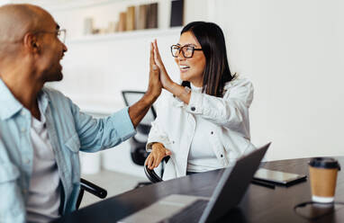 Happy business people sitting at a table and giving each other a high five. Young business people celebrating success in an office. - JLPSF28691