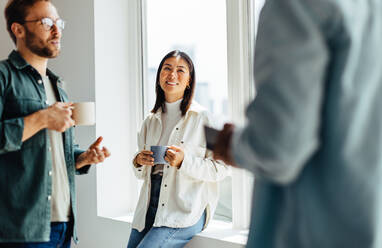 Young business people chatting over a coffee break in an office. Team of young professionals standing together in a modern workplace. - JLPSF28700