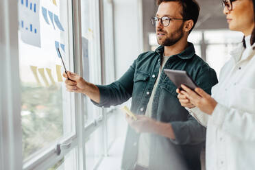 Creative business people brainstorming with sticky notes in an office. Business professionals standing next to a window and discussing their ideas. - JLPSF28706