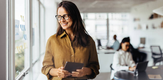 Business woman thinking while holding a tablet pc. Mature sales executive standing in an office with her colleagues in the background. - JLPSF28709
