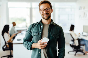 Porträt eines männlichen Berufstätigen, der in einem Büro steht, mit seinen Kollegen im Hintergrund. Ein Geschäftsmann schaut in die Kamera, während er ein Mobiltelefon hält. - JLPSF28728