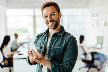 Junger Designer, der ein Mobiltelefon in einem Büro benutzt. Glücklicher Geschäftsmann, der in die Kamera schaut, während er in einem Großraumbüro steht. - JLPSF28730