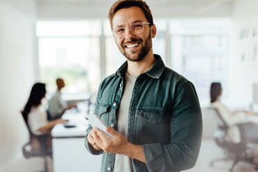 Man smiling at the camera while holding a mobile phone in an office. Business man standing in an open plan office with his colleagues in the background. - JLPSF28731