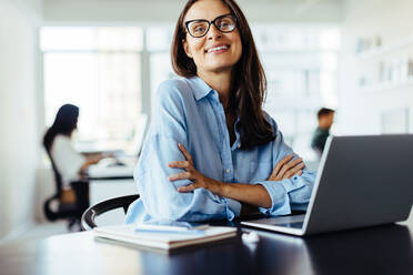 Weibliche Webentwicklerin lächelt in die Kamera, während sie mit einem Laptop in einem Büro sitzt. Reife Geschäftsfrau, die in einem kreativen Büro arbeitet. - JLPSF28738