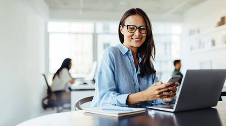 Reife Geschäftsfrau, die ein Mobiltelefon benutzt, während sie in einem Büro mit einem Laptop sitzt. Frau, die in einer Agentur für digitales Marketing arbeitet. - JLPSF28740