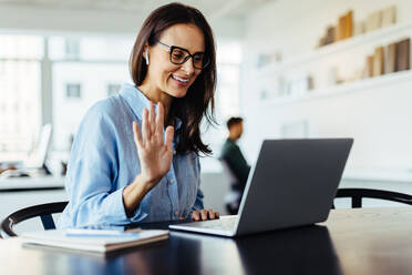 Woman waving on a video call while sitting in an office. Female designer having an online meeting with her business clients on a laptop. - JLPSF28741