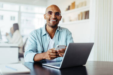 Ein Geschäftsmann lächelt in die Kamera, während er ein Mobiltelefon hält. Ein junger männlicher Designer sitzt in einem Büro mit einem Laptop. - JLPSF28756