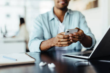 Ein Mann sitzt in einem Büro und benutzt ein Mobiltelefon. Ein Geschäftsmann sendet eine Textnachricht, während er an einem modernen Arbeitsplatz arbeitet. - JLPSF28757
