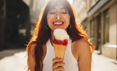 Rothaarige Frau, die ein Eis isst, während sie auf der Straße steht. Glückliche Touristin, die ihren Sommerurlaub in einer neuen Stadt genießt. Bei diesem Foto wurde absichtlich das Korn eines 35-mm-Films verwendet. - JLPPF01541
