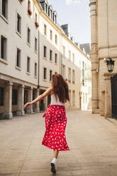 Rearview of a female tourist running through the streets of a city. Woman with red hair exploring and having fun on her summer vacation. This photo has intentional use of 35mm film grain. - JLPPF01543