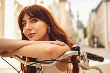 Frau mit roten Haaren, die sich in einer Stadt auf den Lenker eines Fahrrads stützt. Junge Frau, die im Urlaub mit dem Fahrrad unterwegs ist. Bei diesem Foto wurde absichtlich das Korn eines 35-mm-Films verwendet. - JLPPF01544