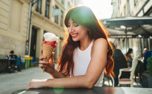 Weibliche Touristin isst ein Eis, während sie in einem Restaurant im Freien sitzt. Glückliche rothaarige Frau, die den Sommerurlaub in einer Stadt genießt. Bei diesem Foto wurde absichtlich das Korn eines 35-mm-Films verwendet. - JLPPF01563