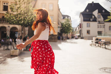 Rückansicht einer fröhlichen Reisenden, die durch die Straßen von Luxemburg läuft. Eine Frau mit roten Haaren, die sich bei der Erkundung Europas im Sommer amüsiert. Dieses Foto weist absichtlich eine 35-mm-Filmkörnung auf. - JLPPF01564