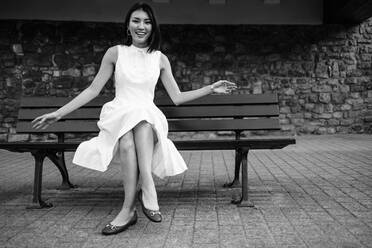 Asiatische Frau lächelt in die Kamera, während sie in einem weißen Kleid auf einer Bank sitzt. Eine Frau, die sich im Freien vergnügt. Bei diesem Foto wurde absichtlich das Korn eines 35-mm-Films verwendet. - JLPPF01567