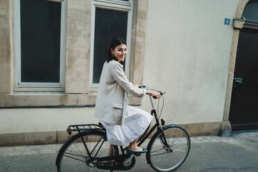 Asiatische Frau, die in die Kamera lächelt, während sie in der Stadt Fahrrad fährt. Eine Frau, die im Urlaub mit dem Fahrrad unterwegs ist. Bei diesem Foto wurde absichtlich das Korn eines 35-mm-Films verwendet. - JLPPF01572