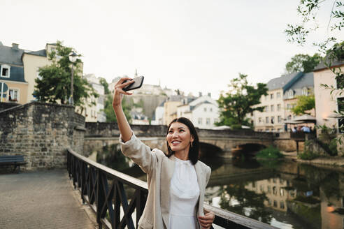 Weibliche Touristin macht ein Selfie neben einem Fluss in Luxemburg-Stadt. Glückliche Frau beim Sightseeing im Urlaub in Europa. Dieses Foto hat absichtlich 35mm Filmkorn verwendet. - JLPPF01579
