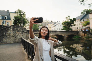 Frau, die während eines Urlaubs in Europa ein Selfie an einem Fluss macht. Weibliche Reisende beim Sightseeing in Luxemburg. Bei diesem Foto wurde absichtlich das Korn eines 35-mm-Films verwendet. - JLPPF01580