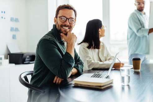 Ein Geschäftsmann lächelt in die Kamera, während er in einem Sitzungssaal sitzt. Ein glücklicher Geschäftsmann sitzt in einer Präsentation mit einem Laptop. - JLPSF28766