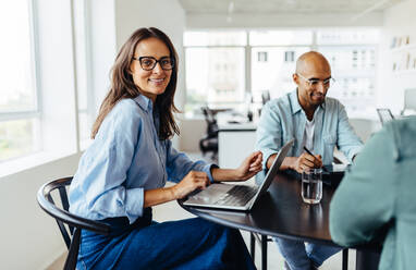 Mature female designer sitting around a table and smiling at the camera. Happy business woman having a meeting with her team in an office. - JLPSF28769