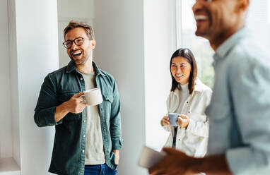 Geschäftsleute, die während einer Kaffeepause bei der Arbeit zusammen lachen. Eine Gruppe junger Berufstätiger steht in einem Büro zusammen. - JLPSF28778