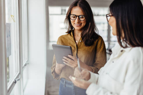 Geschäftsfrauen, die an einem Fenster stehen und ihre Ideen besprechen. Weibliche Geschäftsleute beim Brainstorming mit Notizen und einem Tablet. - JLPSF28782