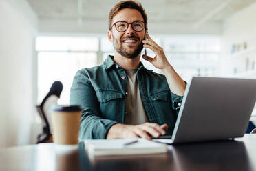 Entrepreneur making a phone call while working on a laptop. Young business man making plans with a client in an office. - JLPSF28794