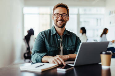 Porträt eines Geschäftsmannes, der in einem Büro sitzt, mit seinen Kollegen im Hintergrund. Glücklicher Geschäftsmann, der in einem Co-Working-Büro arbeitet. - JLPSF28798