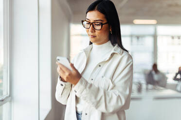 Asiatische Geschäftsfrau, die an ihrem Arbeitsplatz ein Mobiltelefon benutzt. Weibliche Geschäftsfrau, die in einem Büro stehend ihre Textnachrichten durchsieht. - JLPSF28801