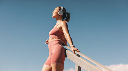Frau in Sportkleidung hört Musik über Kopfhörer im Freien. Weibliche Athletin macht eine Pause vom Training. Kaukasische Sportlerin steht gegen den Himmel. - JLPSF28809