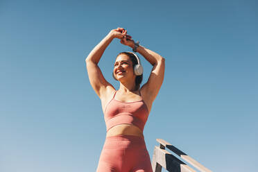 Woman in sportswear stretching her arms outdoors. Happy sportswoman doing a warm up exercise with headphones on. Female athlete working out against the sky. - JLPSF28810