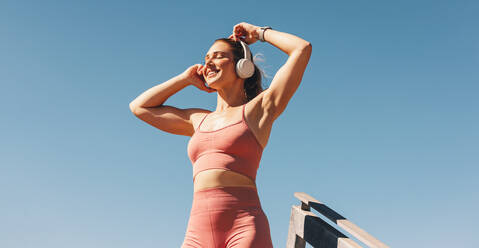 Caucasian sports woman listening to music on headphones outdoors. Woman in sportswear standing against the sky. Happy female athlete taking a break from her workout. - JLPSF28811