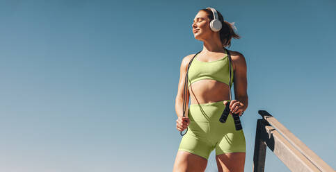 Sportliche Frau, die Musik hört und ein Springseil hält. Frau in Sportkleidung, die im Freien vor dem Himmel steht. Sportlerin, die eine Pause von ihrem Kardio-Training macht. - JLPSF28812