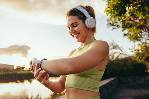 Sportliche Frau, die mit einer Smartwatch im Freien eine Fitness-Wiedergabeliste auswählt. Glückliche Frau beim morgendlichen Lauf mit Kopfhörern. Frau, die Technologie für ihr Workout nutzt. - JLPSF28814