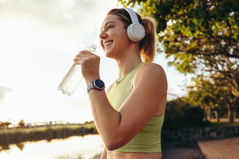 Sportliche Frau, die im Freien Wasser aus einer Flasche trinkt. Glückliche Frau, die eine Pause von ihrem morgendlichen Training macht. Sportliche Frau, die neben einem See steht. - JLPSF28816