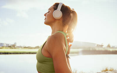 Woman in her 30s prepares to start her exercise routine near a peaceful lake. She has her headphones in and is wearing sportswear while stretching and warming up. Her goal is to dedicate her time to her wellbeing and health, while also creating a healthy lifestyle for herself. - JLPSF28818