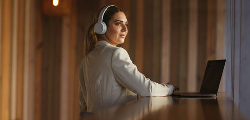 Berufstätige Frau mit drahtlosen Kopfhörern in einem Coworking Space. Geschäftsfrau, die Musik hört, während sie an einem Laptop arbeitet. Frau, die aus der Ferne arbeitet. - JLPSF28837