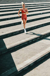 Female runner exercising down the stairs outdoors. Sporty woman doing a cardio exercise in the morning. Happy caucasian woman working out in sportswear. - JLPSF28849