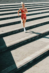 Weibliche Läuferin, die im Freien die Treppe hinunter trainiert. Sportliche Frau, die morgens ein Ausdauertraining macht. Glückliche kaukasische Frau, die in Sportkleidung trainiert. - JLPSF28849
