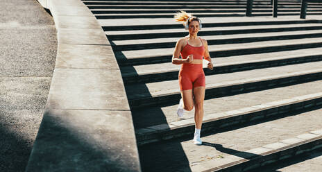 Eine Läuferin verbrennt beim Ausdauertraining Kalorien. Eine sportliche Frau joggt im Freien die Treppe hinunter. Eine Frau trainiert mit Kopfhörern am Morgen. - JLPSF28850