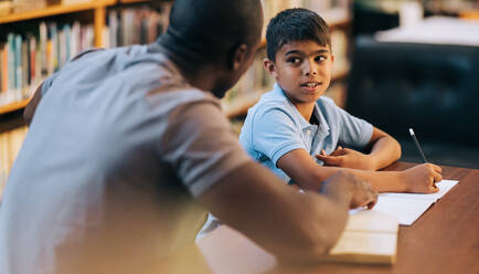 Nachhilfeunterricht und akademische Unterstützung in der Grundschule. Kleiner Schüler im Gespräch mit seinem Lehrer während einer Nachhilfestunde in einer Bibliothek. Grundschüler sitzt mit einem Pädagogen zusammen. - JLPSF28867