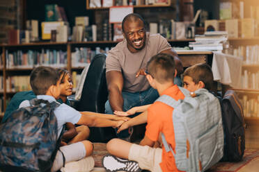 Male school teacher huddling with his students in a library. Man teaching a group of primary school children. Motivation and mentorship in elementary school. - JLPSF28870