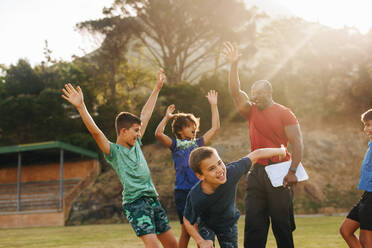 Grundschulkinder feiern mit ihrem Sportlehrer beim Sportunterricht. Männlicher Trainer motiviert eine Gruppe von Grundschulkindern. Sport und Freizeit in der Schule. - JLPSF28897