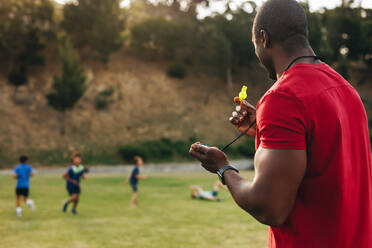 Ein Trainer hält eine Trillerpfeife während einer Sportstunde mit Schulkindern. Ein Mann trainiert eine Gruppe von Grundschulkindern auf einem Schulgelände. Sport und Erholung in der Schule. - JLPSF28901